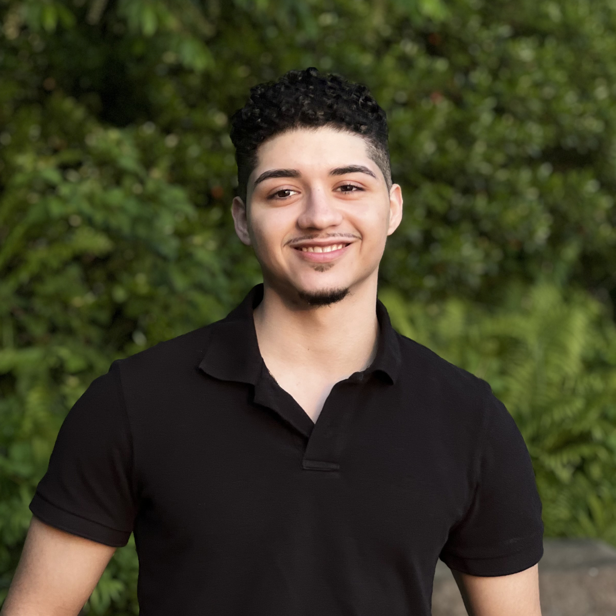 A portrait image of me smiling in a black polo shirt. Behind me is a wall of grass.
